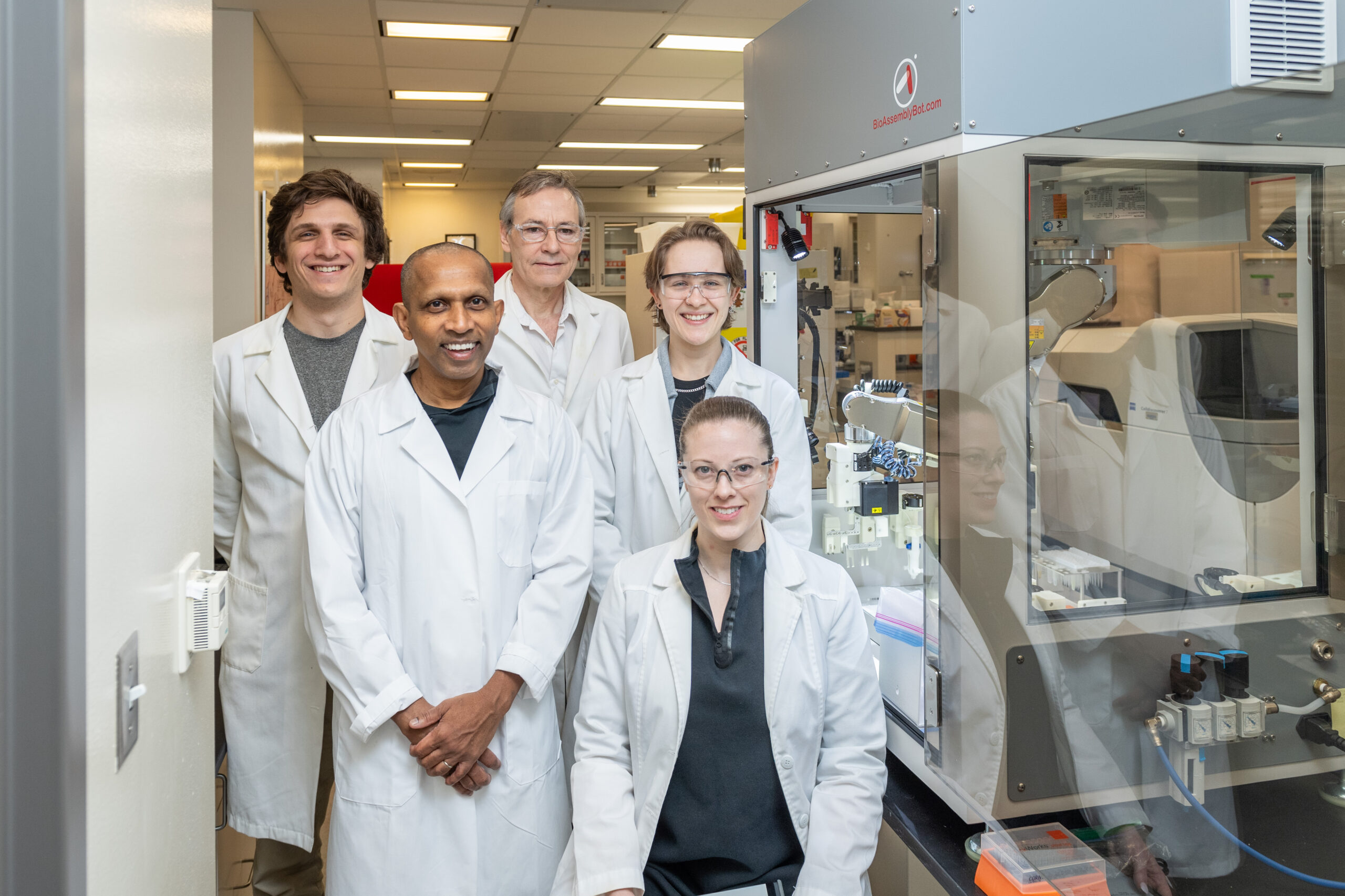 Lab members stand in white lab coats, smiling next to a robotic 3D printer in the lab.